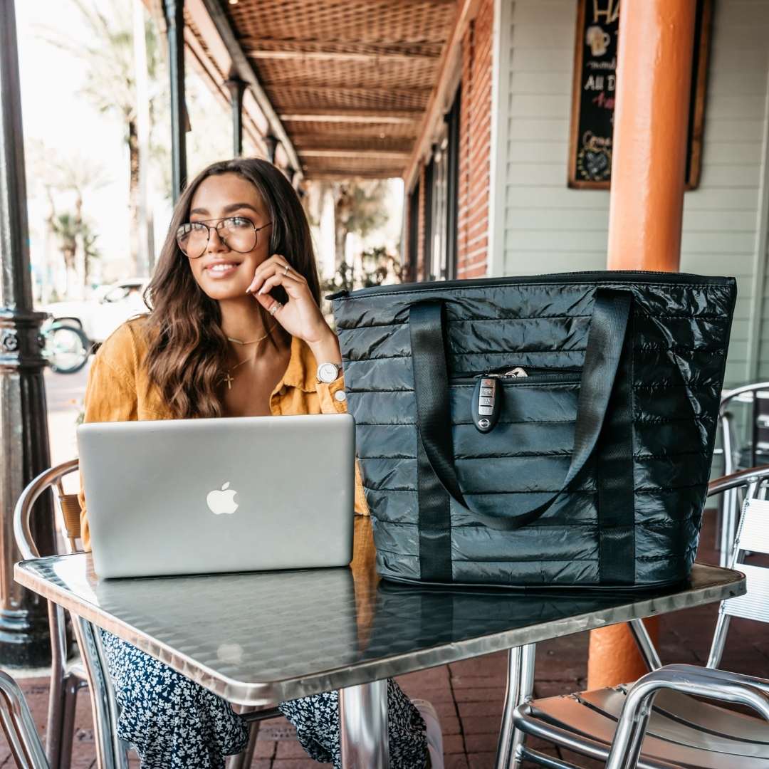 Woman sitting at an outdoor cafe with a laptop and a large shimmer black puffer tote on the table.