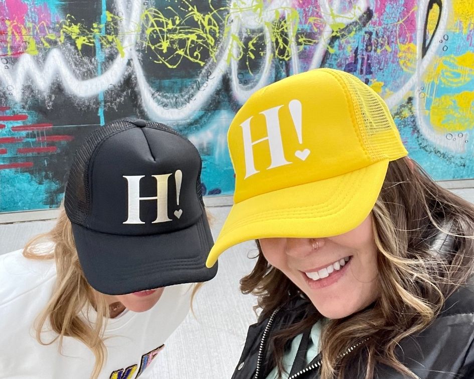 Two people wearing black and yellow hats with 'H!' on them in front of a colorful graffiti wall.
