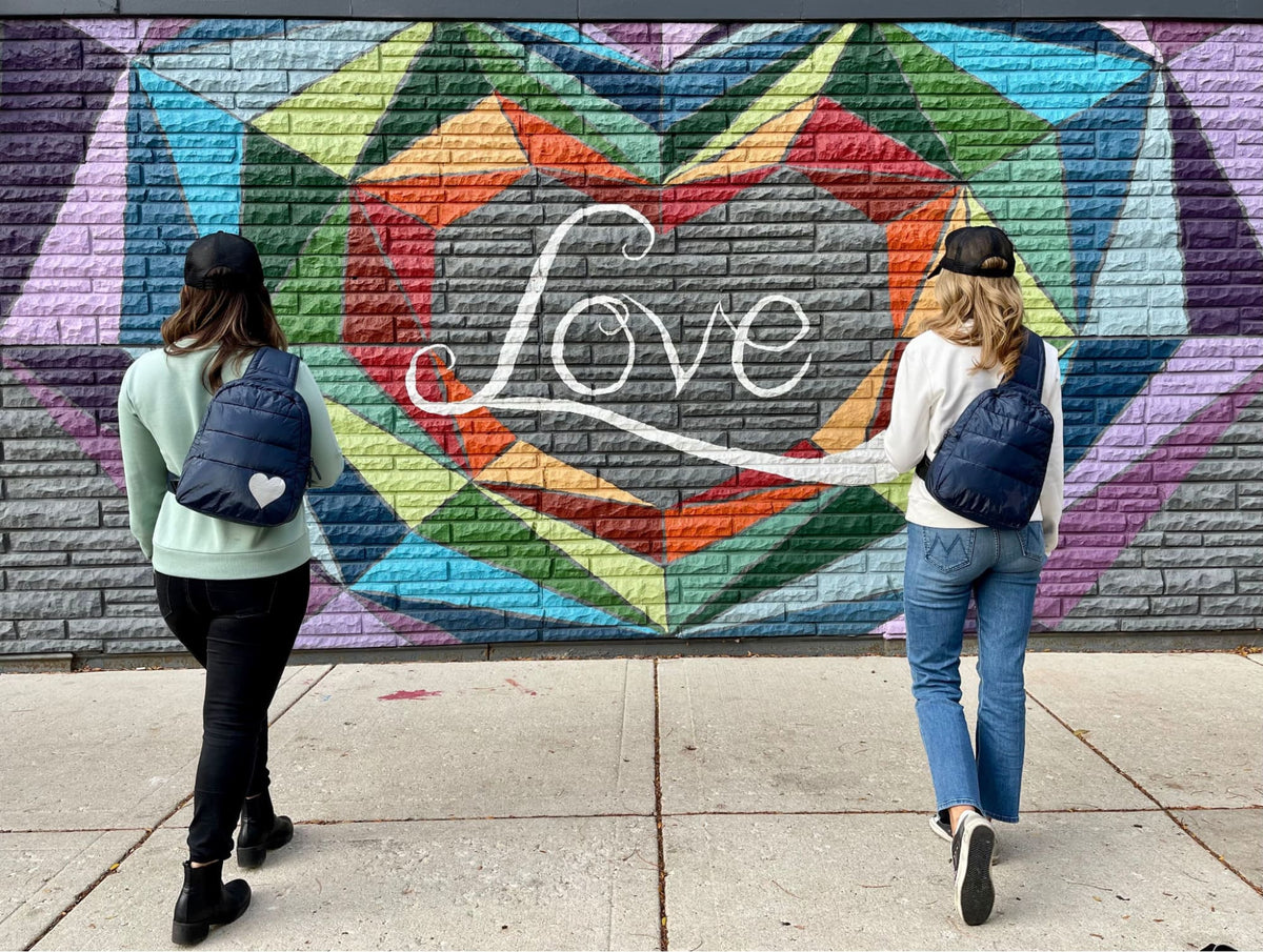 Two women wearing Hi Love Travel puffer backpacks in mint green and navy in front of a colorful "Love" mural.