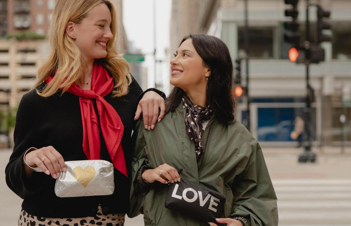 Two women standing on a city street, each holding a small clutch bag. The woman on the left wears a black top and a bright red scarf, holding a silver clutch with a gold heart.