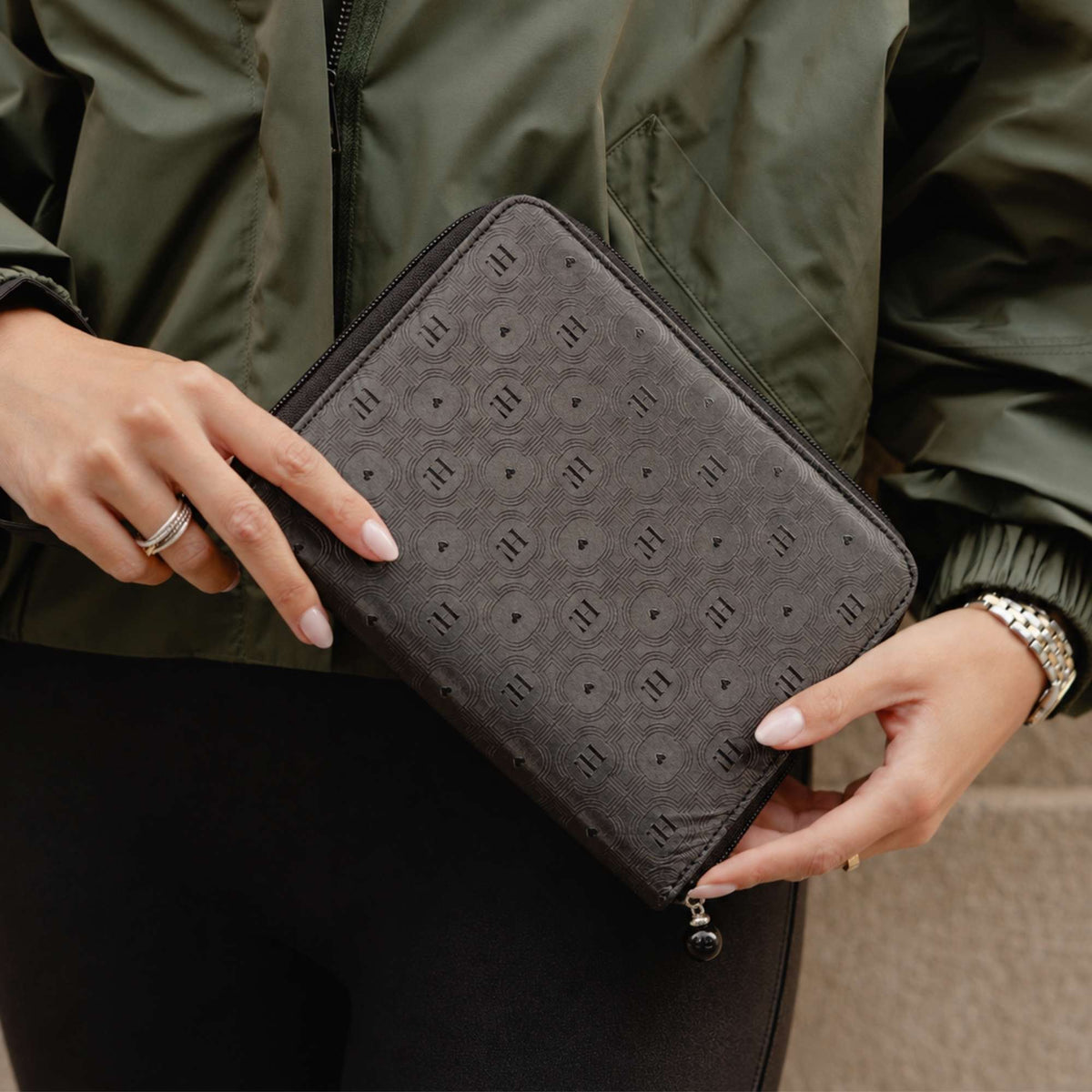 A person holding a black zippered jewelry case, with an embossed geometric pattern, wearing an olive green jacket and black pants.