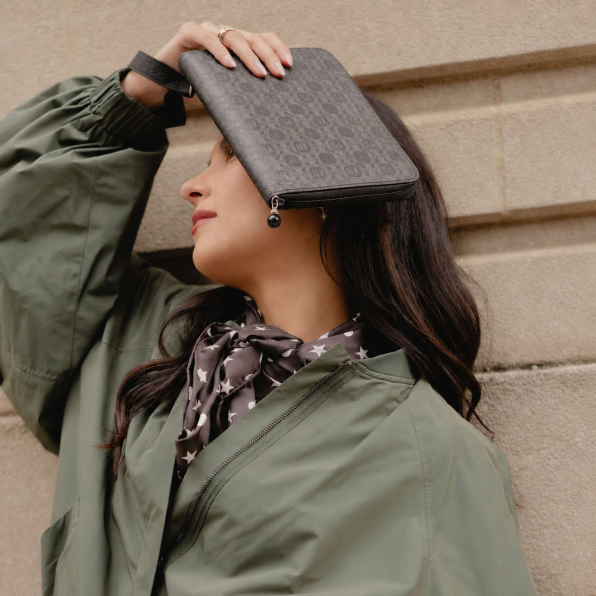 A woman wearing an olive green jacket and a patterned scarf holds a black monogram jewelry case against her forehead, posing in front of a beige stone wall.