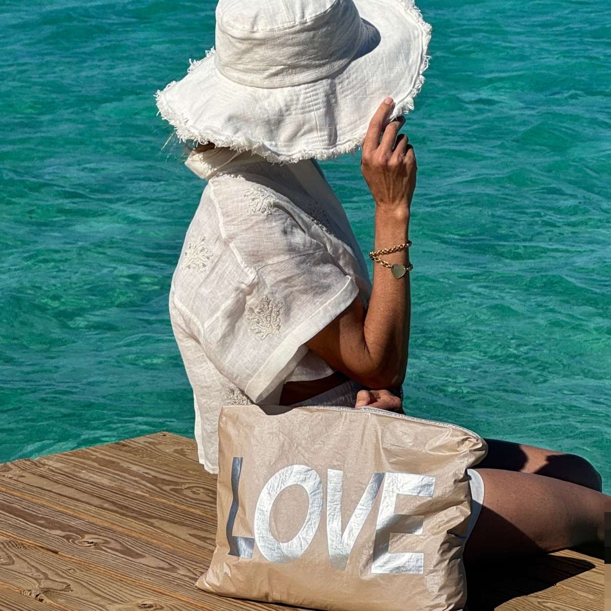 Person sitting on a dock by the water, wearing a white hat and dress, with a 'LOVE' bag.