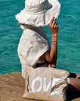 Person sitting on a dock by the water, wearing a white hat and dress, with a 'LOVE' bag.