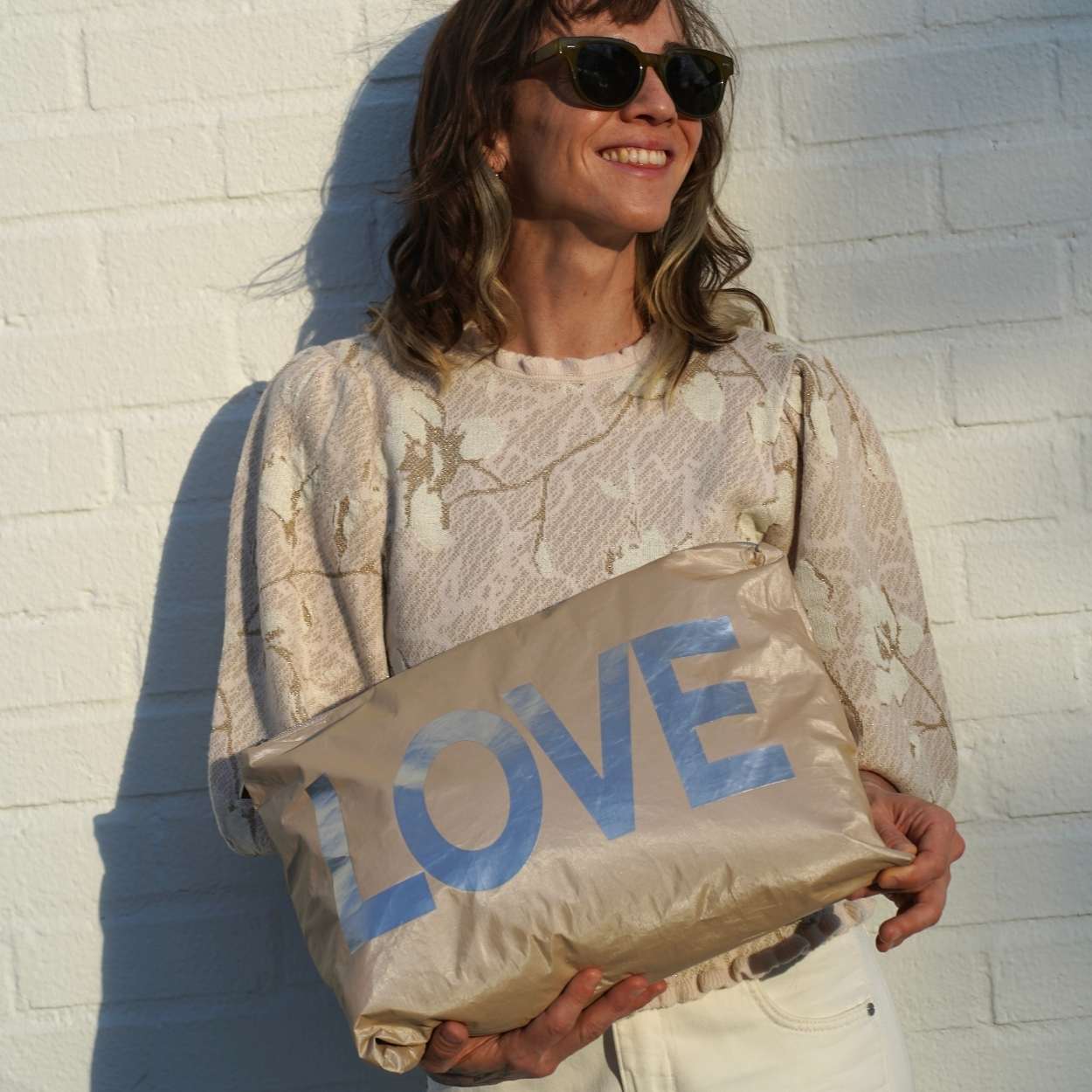 Person holding a beige bag with 'LOVE' printed on it against a white brick wall.