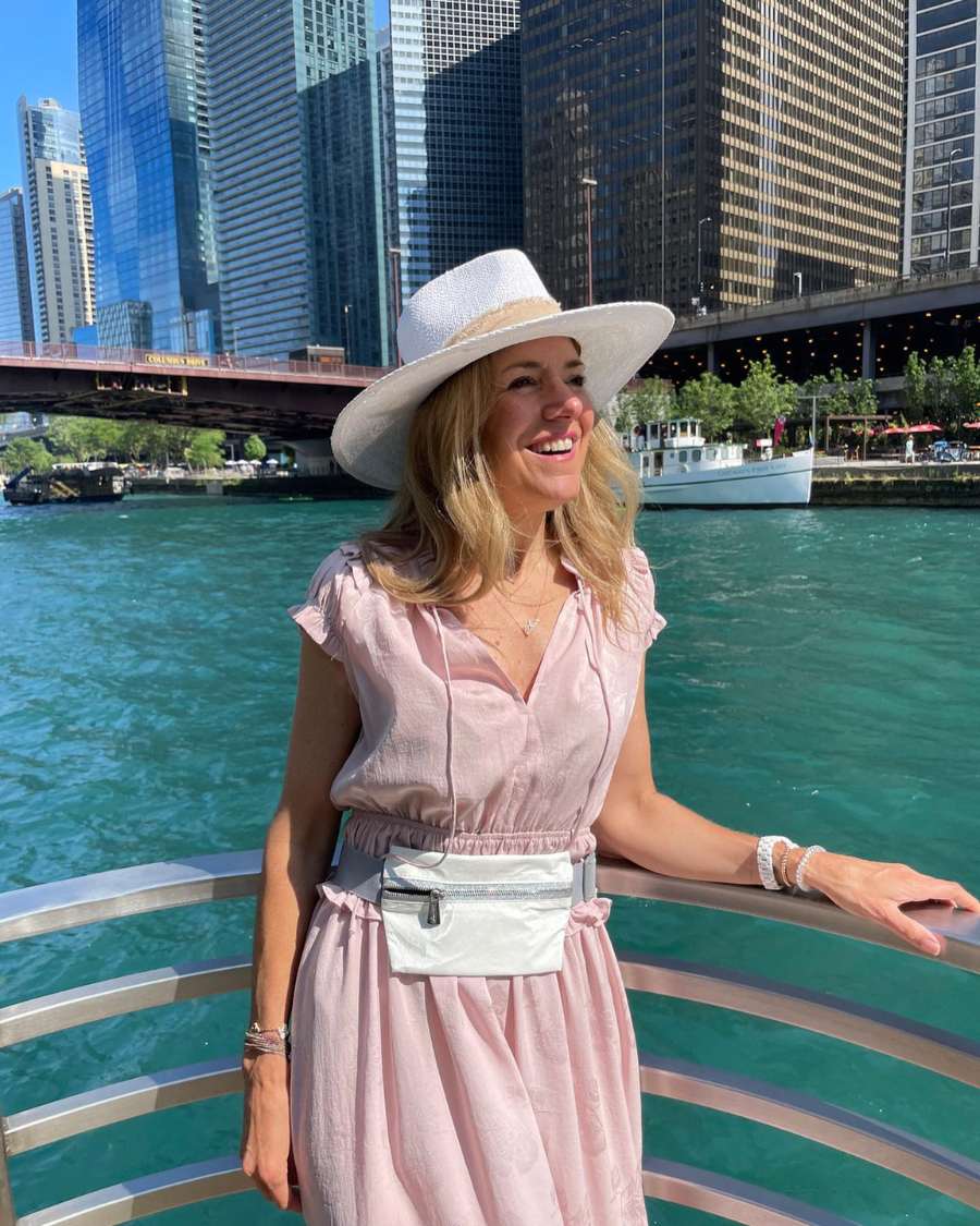 Woman in a pink dress and white hat standing by a waterfront with city skyline in the background