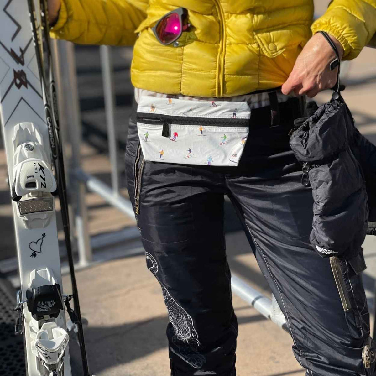 Person in black snow pants, yellow jacket, ski patterned fanny pack, and gloves holding skis in an outdoor setting