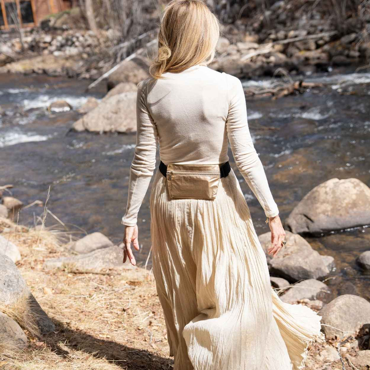 A woman stands outdoors by a flowing river, shown from the back. She is wearing a fitted beige mock-neck long-sleeve top, a matching pleated maxi skirt, and a shiny metallic gold belt bag with a black strap.