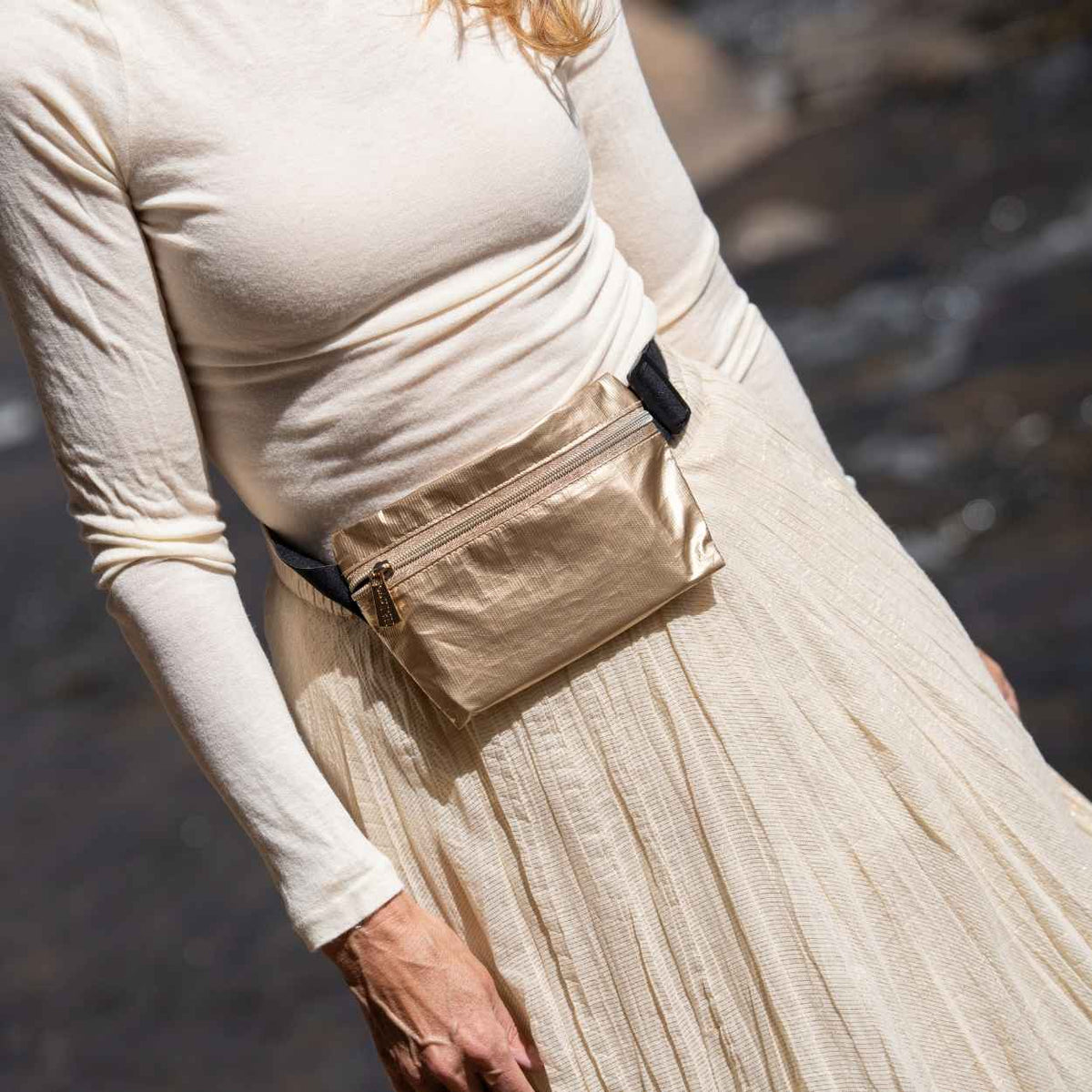 Person in cream long sleeve and pleated skirt with gold fanny pack around the waist in blurred background. 