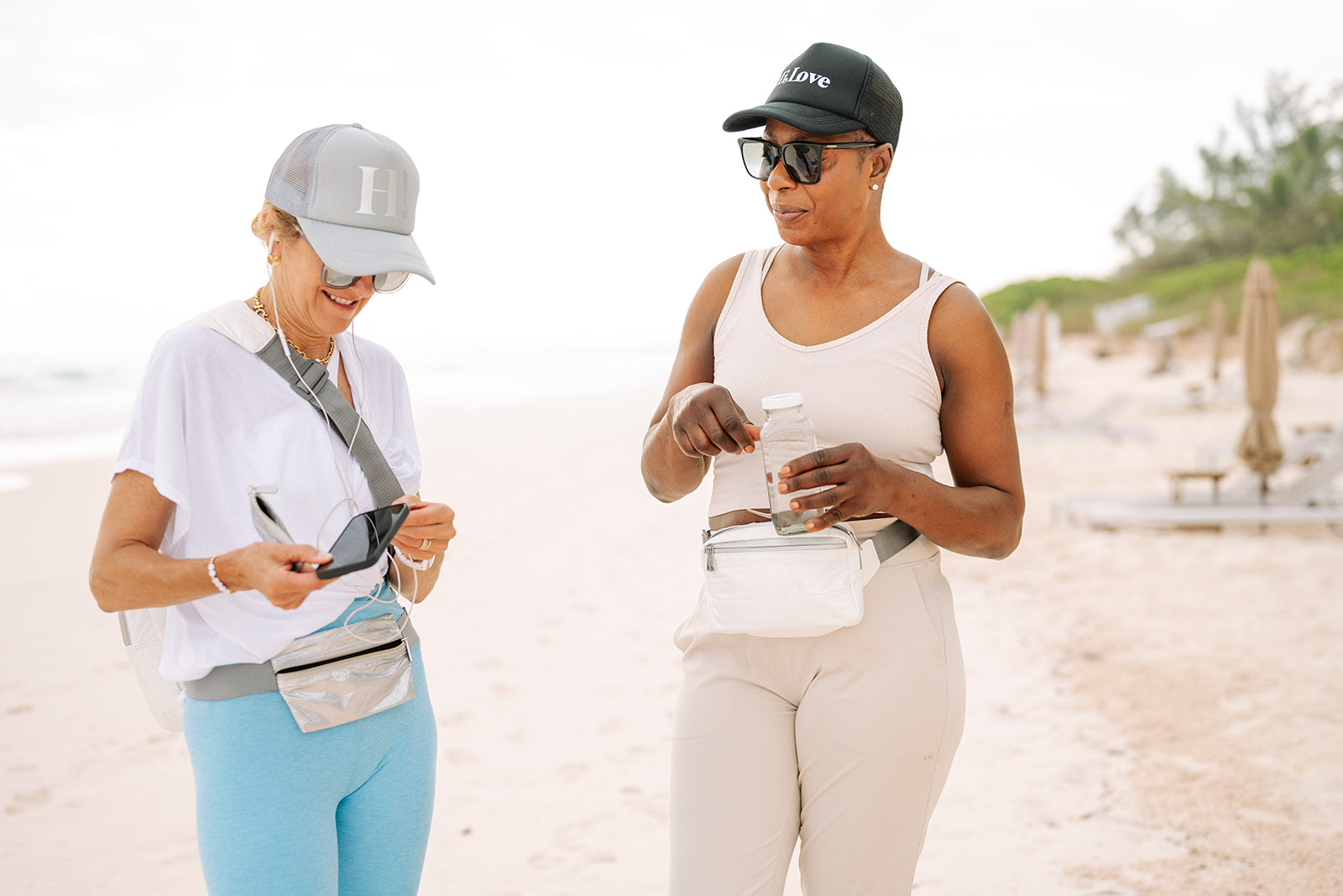 Two women walking on a beach, one using a phone and the other holding a water bottle, both wearing fanny pack belt bags.