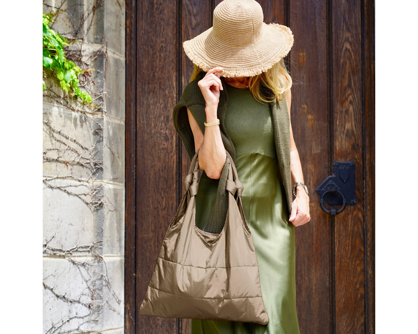 Woman standing in front of wooden door in green dress, straw sunhat, and shimmer bronze love me knot tote bag