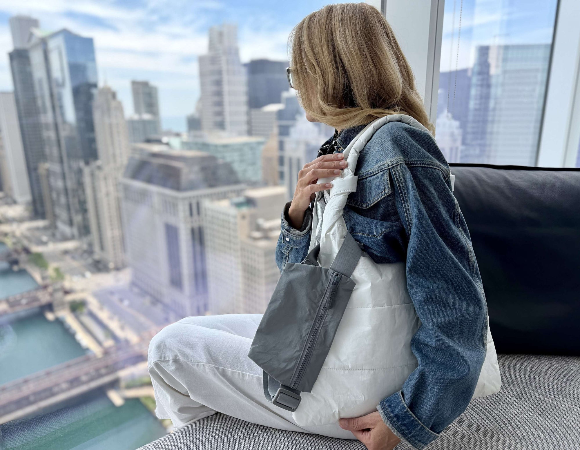 Person sitting on a couch with a cityscape view, holding a white tote bag with gray belt bag clipped on it.
