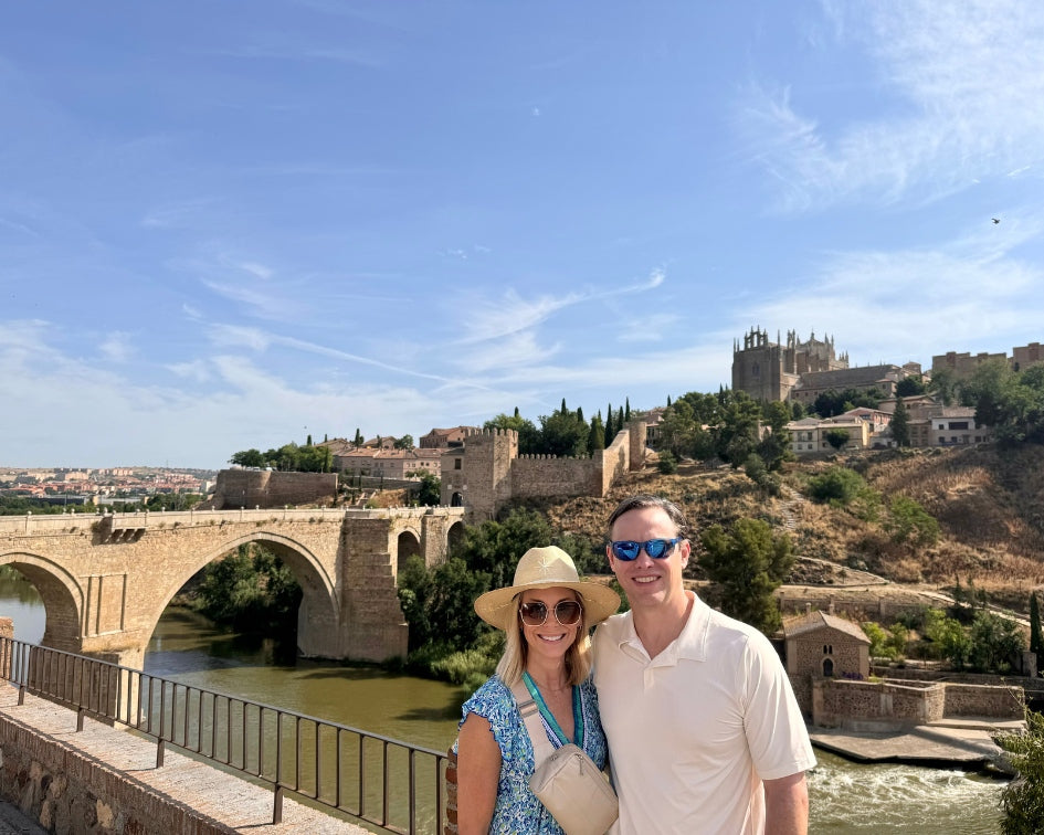 Couple standing in front of a historic bridge and castle under a clear blue sky.