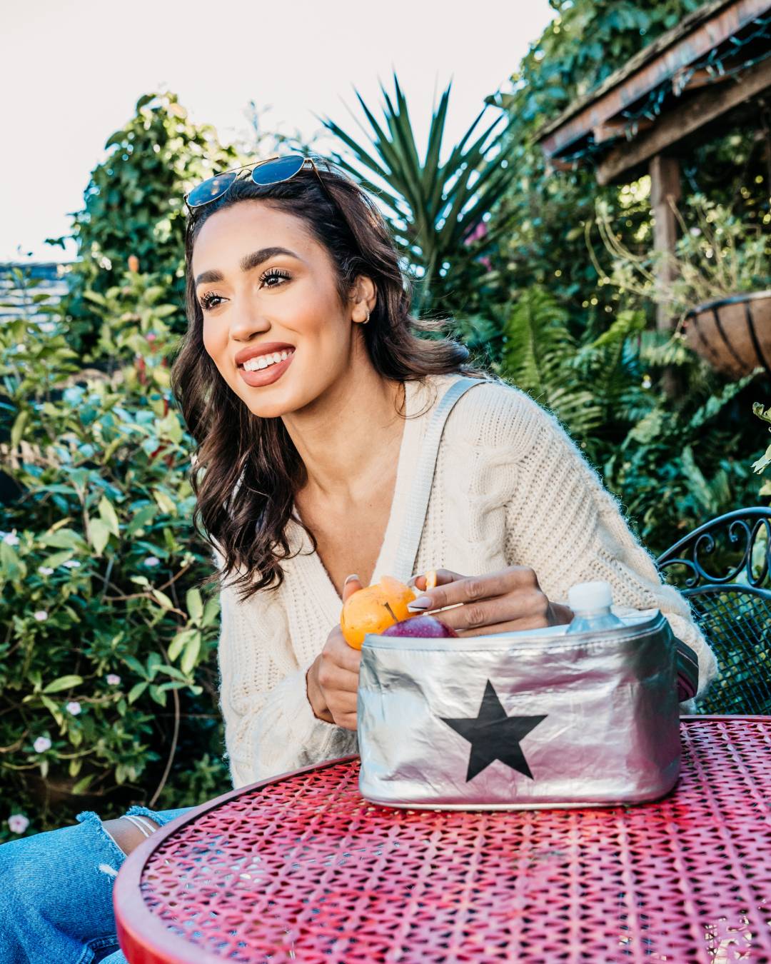 A woman sitting at a red metal patio table outdoors, holding a shiny silver insulated bag with a large black star on the front. She is wearing a cream knit sweater and blue jeans, with green garden foliage in the background.