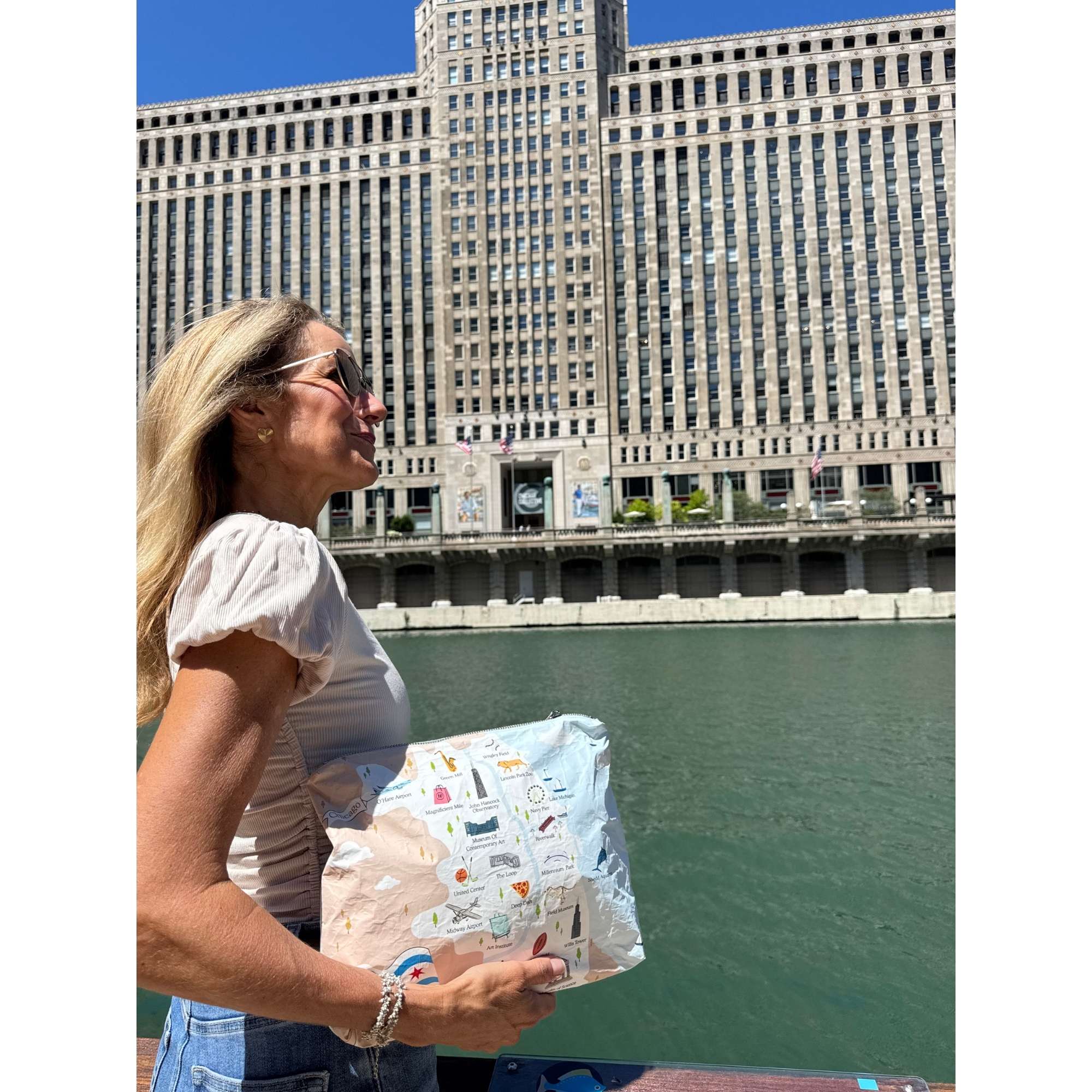 A woman wearing sunglasses and a white top holding a light beige tote bag and Chicago patterned pouch. The background shows a classical stone building, a modern skyscraper, and a clear blue sky.