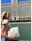 A woman wearing sunglasses and a white top holding a light beige tote bag and Chicago patterned pouch. The background shows a classical stone building, a modern skyscraper, and a clear blue sky.