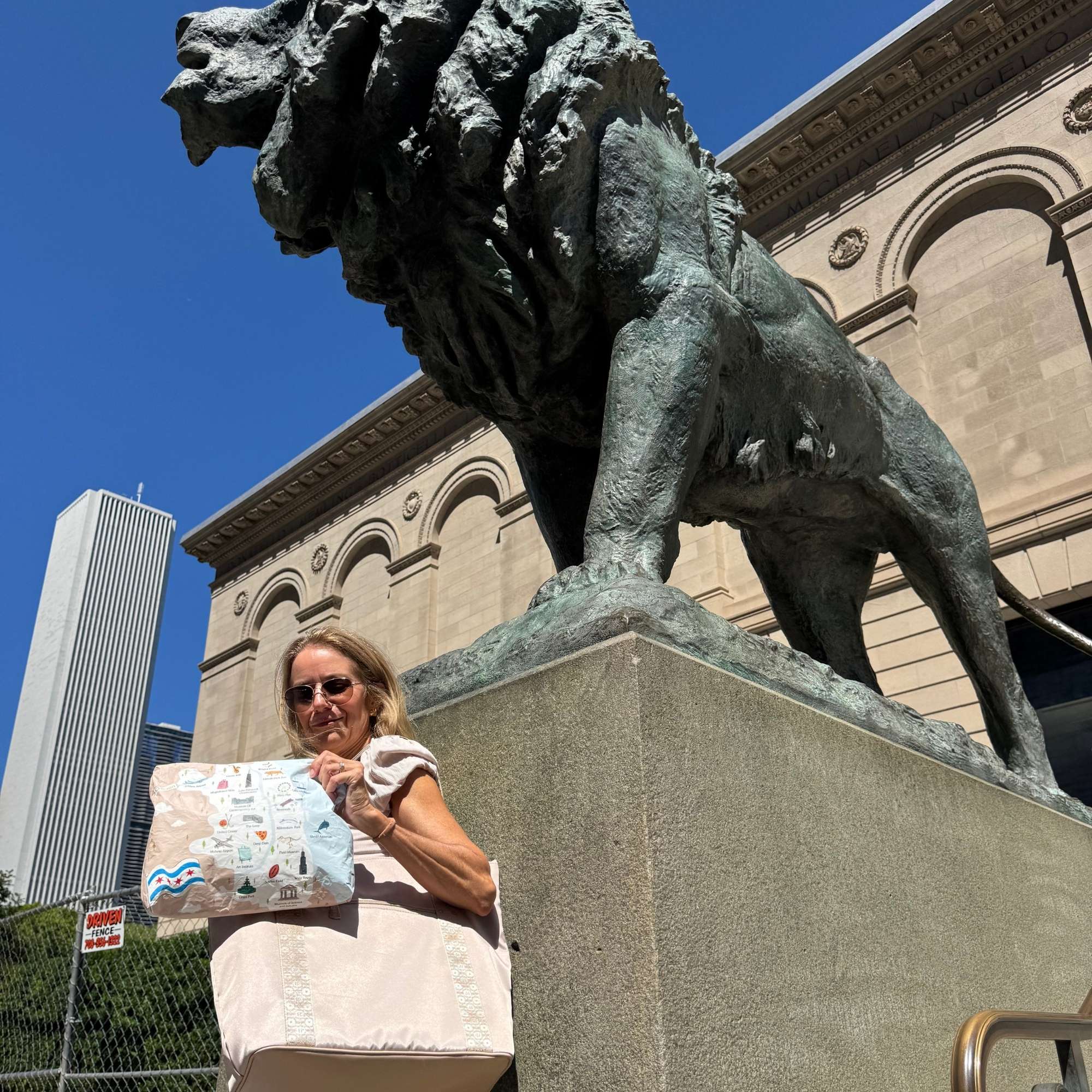 A woman wearing sunglasses and a white top stands beside a large bronze lion statue on a stone pedestal, holding a light beige tote bag and Chicago patterned pouch. The background shows a classical stone building, a modern skyscraper, and a clear blue sky.