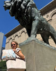 A woman wearing sunglasses and a white top stands beside a large bronze lion statue on a stone pedestal, holding a light beige tote bag and Chicago patterned pouch. The background shows a classical stone building, a modern skyscraper, and a clear blue sky.