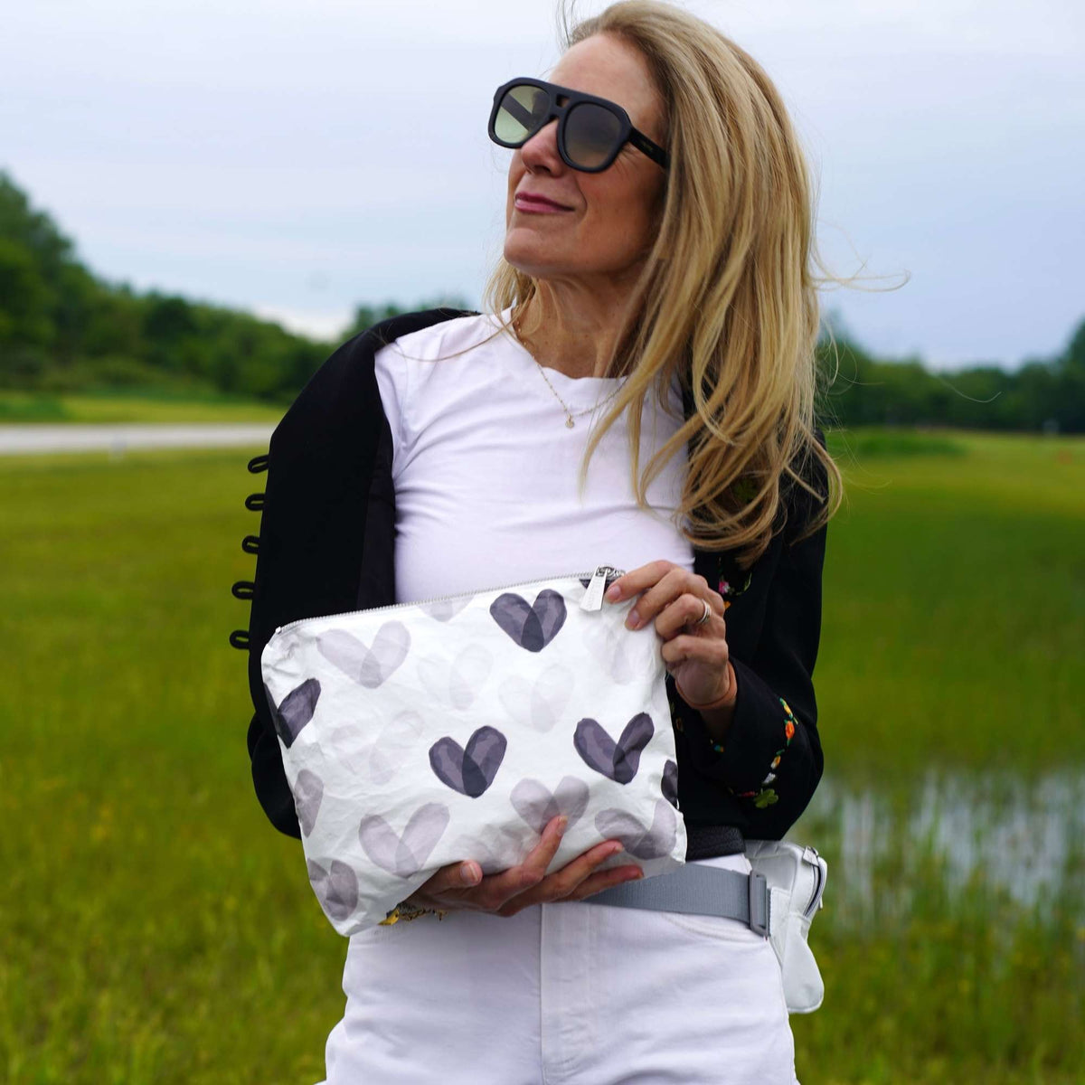 A woman outdoors in a grassy field wearing black sunglasses, a white t-shirt, a black cardigan, and white pants, holding a black and gray watercolor hearts patterned pouch.