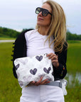 A woman outdoors in a grassy field wearing black sunglasses, a white t-shirt, a black cardigan, and white pants, holding a black and gray watercolor hearts patterned pouch.