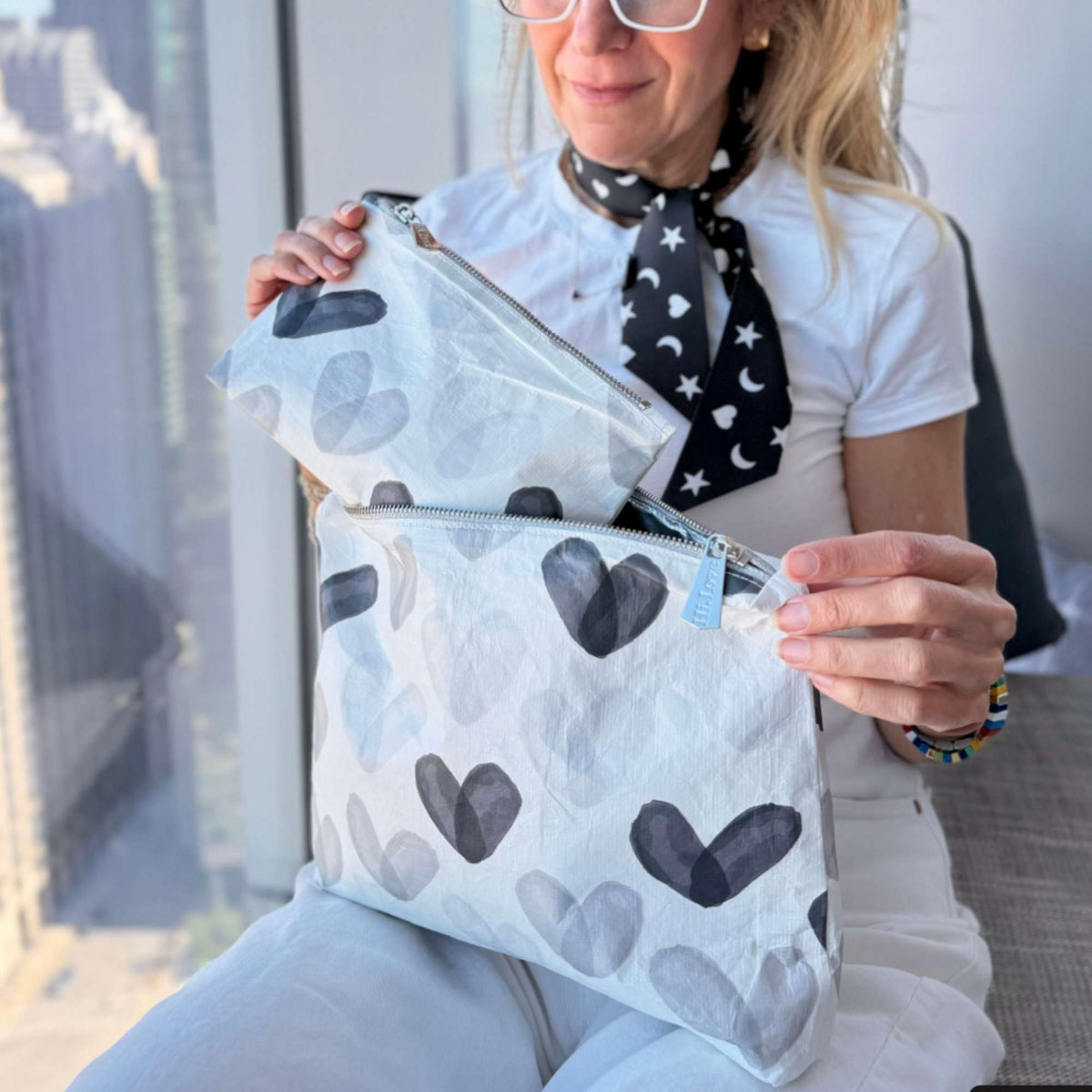A woman holds two zip pouches with a watercolor heart pattern in white, gray, and black, set against a bright, modern indoor background.