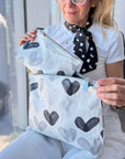A woman holds two zip pouches with a watercolor heart pattern in white, gray, and black, set against a bright, modern indoor background.