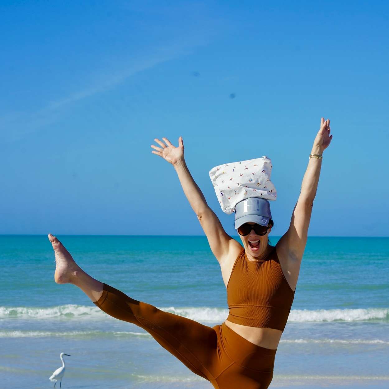 Person in brown outfit with a hat and pouch on their head, standing on one leg on a beach with blue sky and ocean.