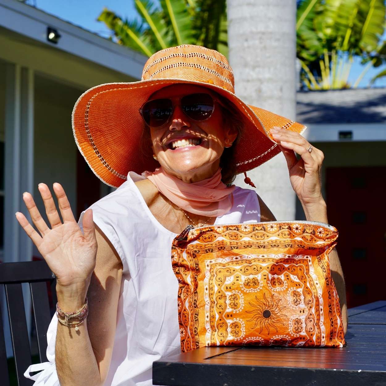 Woman wearing an orange sun hat and sunglasses, holding a matching orange bandana pouch, outdoors.
