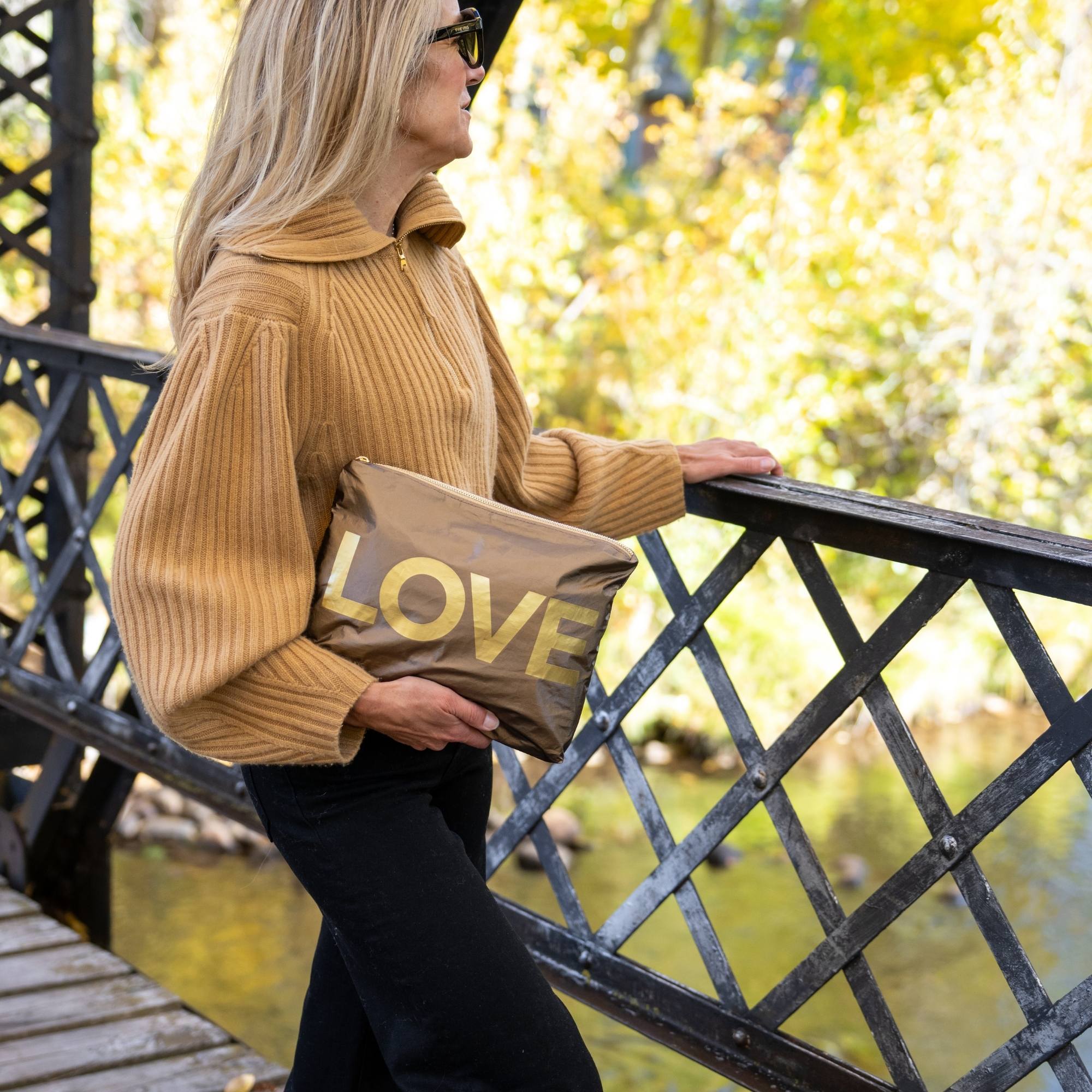Woman holding a bronze metallic medium zipper pouch with oversized gold LOVE lettering while standing on a bridge in a fall setting.
