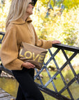 Woman holding a bronze metallic medium zipper pouch with oversized gold LOVE lettering while standing on a bridge in a fall setting.