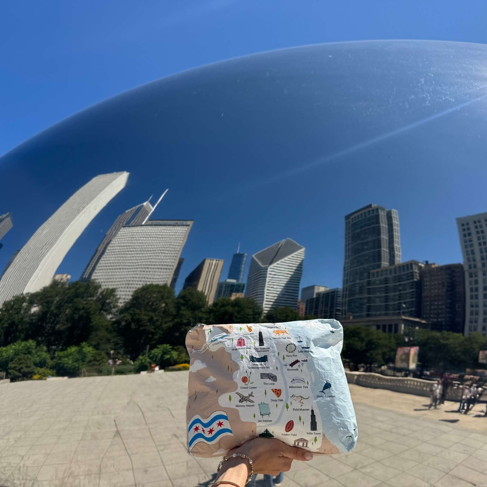 A person holds up a Chicago patterned pouch in front of a wide city plaza with the Chicago Bean sculpture and skyscrapers under a bright blue sky.