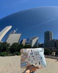 A person holds up a Chicago patterned pouch in front of a wide city plaza with the Chicago Bean sculpture and skyscrapers under a bright blue sky.
