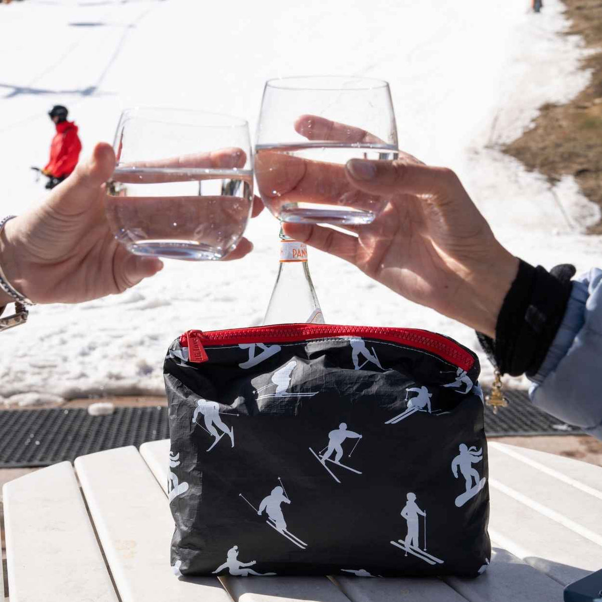 Two hands clinking glasses outdoors in a snowy setting with black pouch on table featuring ski and snowboarder pattern