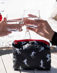 Two hands clinking glasses outdoors in a snowy setting with black pouch on table featuring ski and snowboarder pattern