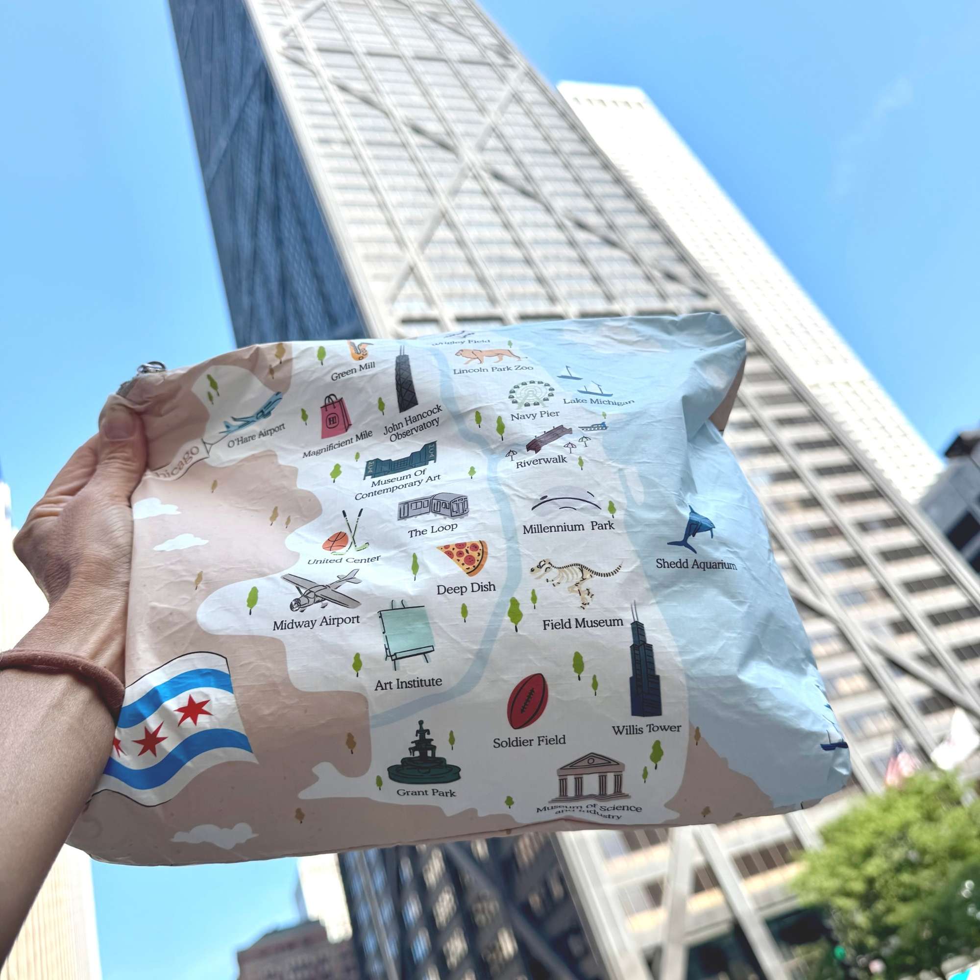 A hand holds a Chicago printed pouch with illustrated landmarks and labels, including Chicago Museum Art Institute, Field Museum, and others, against a real outdoor city background with tall buildings and blue sky.