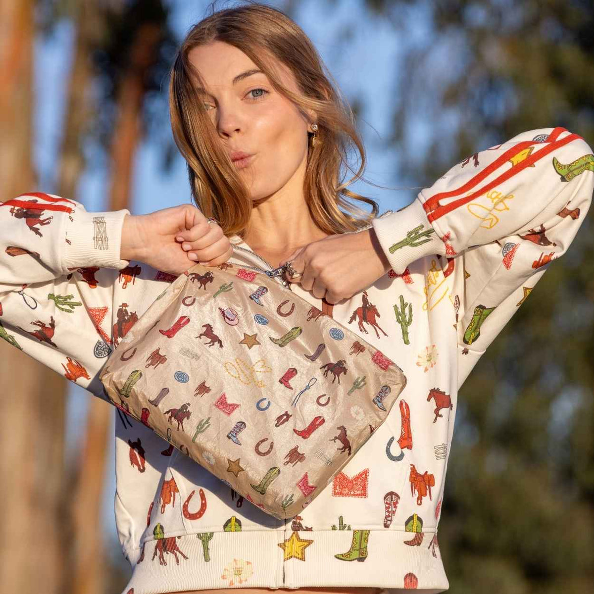 Girl holding a beige pouch with western theme icons printed wearing the coordinating sweatshirt in an outdoor setting.