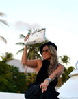Woman in black dress and hat holding a beige clutch with palm trees in the background