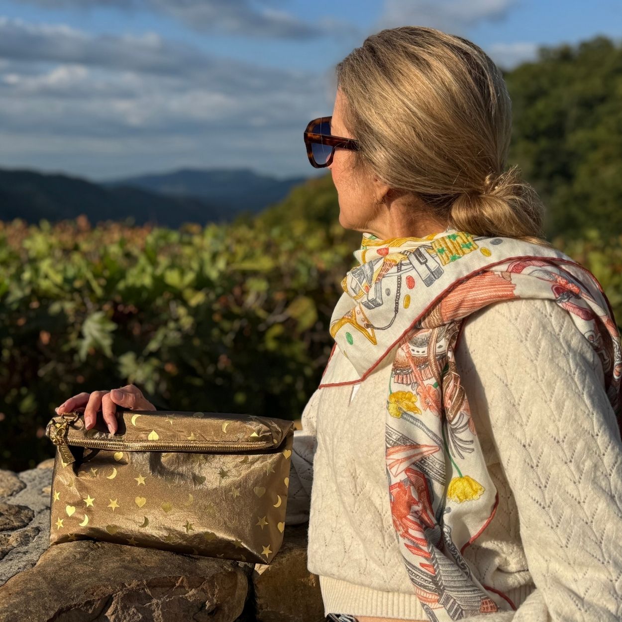 A woman in profile wearing sunglasses, a cream cable-knit sweater, and a colorful patterned scarf, holding a metallic bronze clutch with gold moon and star designs against a scenic outdoor background of mountains and greenery.