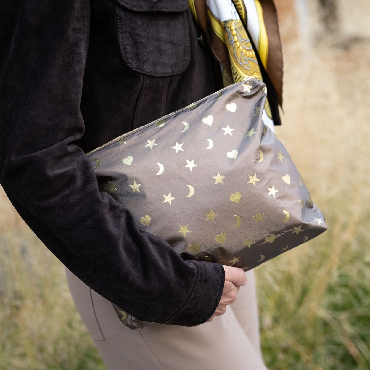 A person outdoors holding a bronze zippered pouch decorated with metallic gold stars, moons, and hearts. The person is wearing a dark brown suede jacket and beige pants.