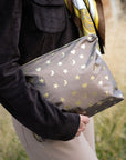 A person outdoors holding a bronze zippered pouch decorated with metallic gold stars, moons, and hearts. The person is wearing a dark brown suede jacket and beige pants.