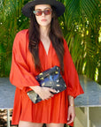 Woman in a red dress and black hat holding a clutch with a butterfly pattern, standing outdoors.
