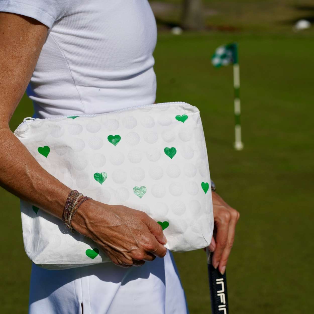 Person in all white outfit holding white bag with green hearts and golf club on golf course