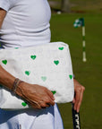 Person in all white outfit holding white bag with green hearts and golf club on golf course