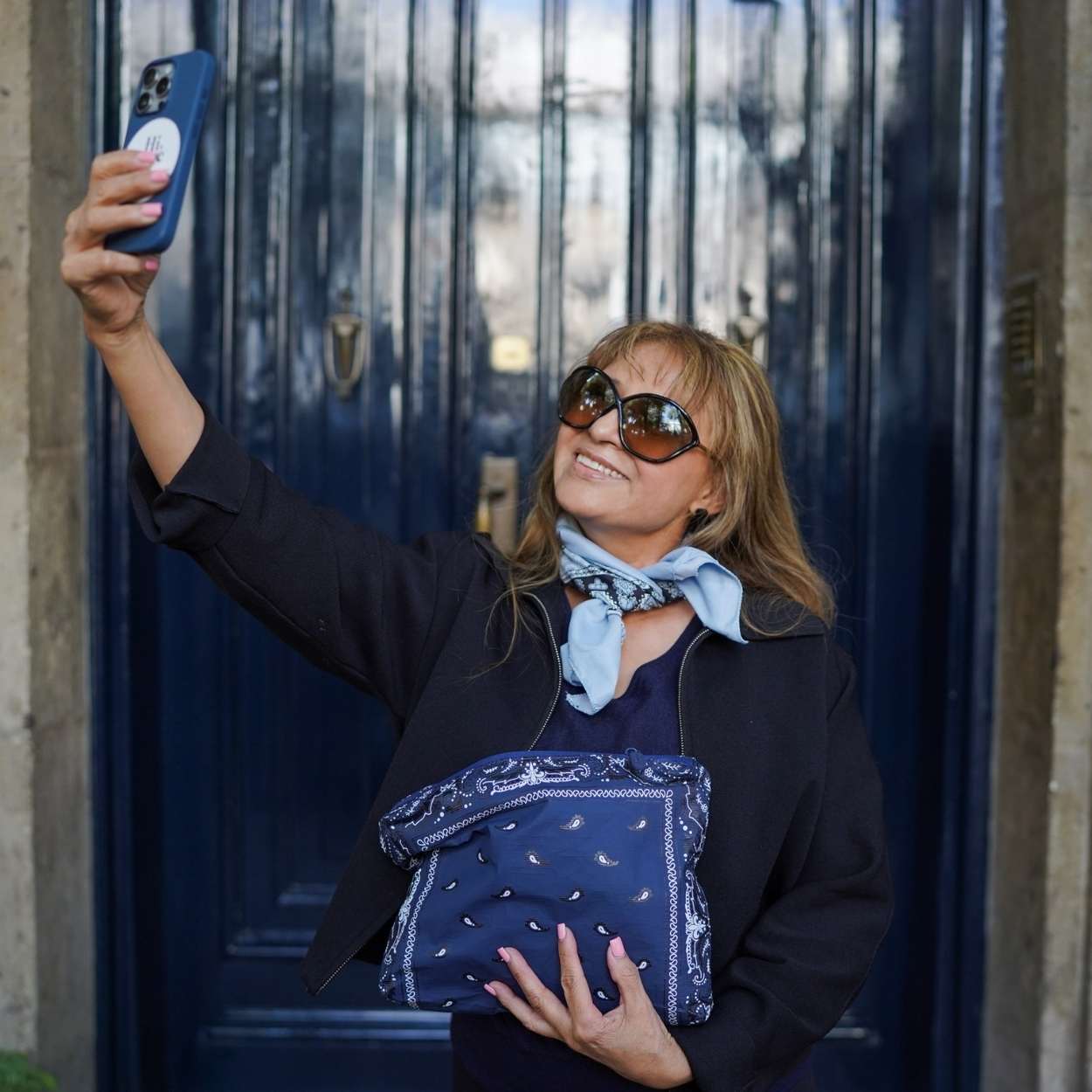 Woman taking a selfie with a blue phone case, wearing sunglasses and holding a navy bandana pouch.
