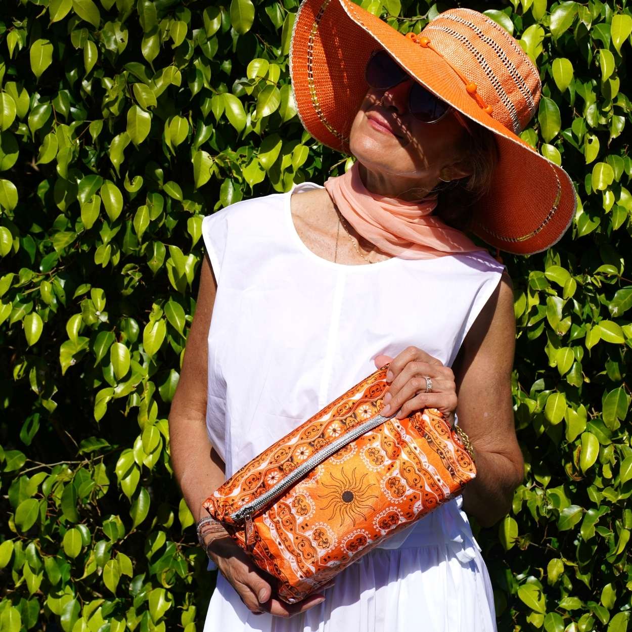 Woman wearing an orange hat and holding an orange bandana patterned bag against a green leafy background