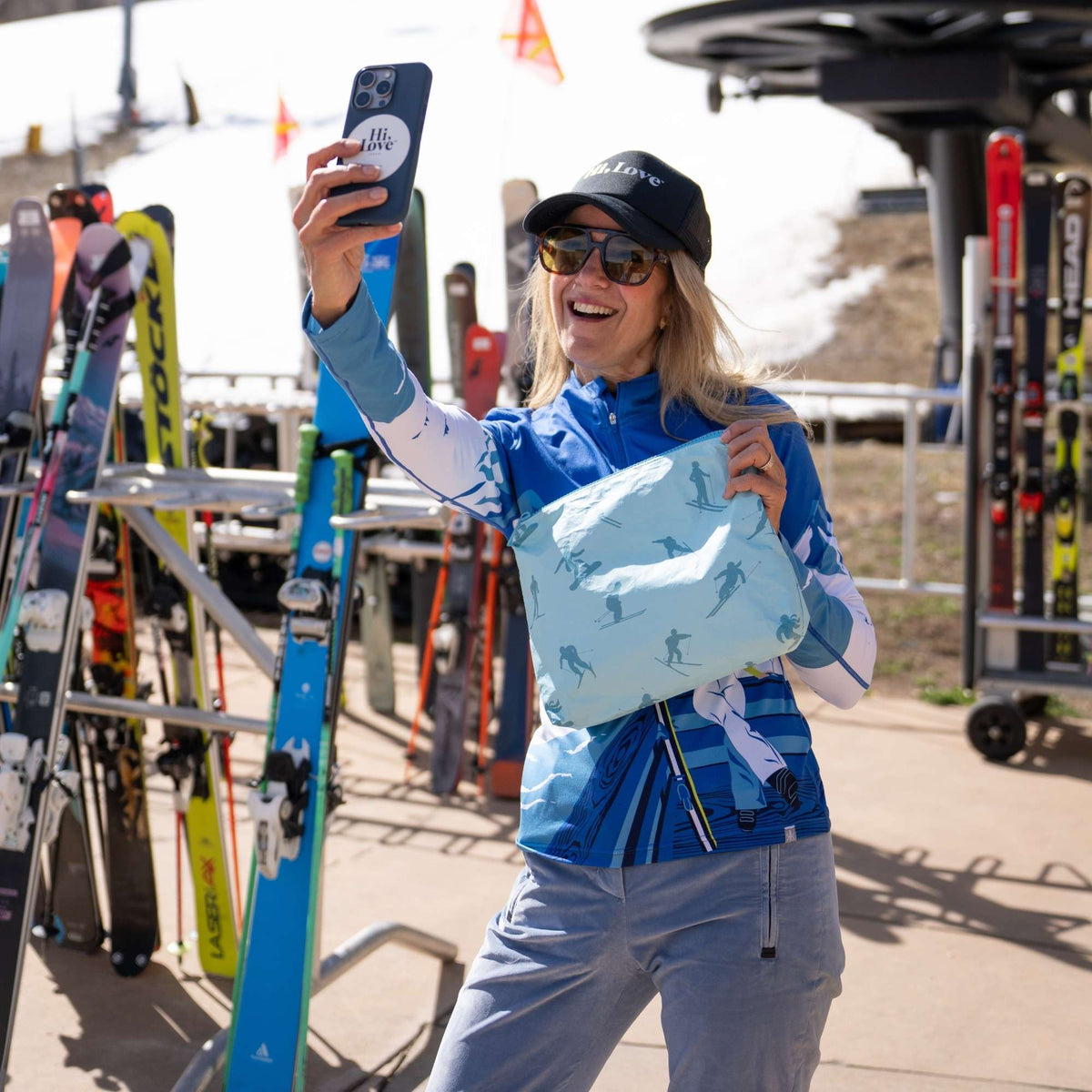 Woman taking a selfie at a ski resort while holding a light blue on blue medium zipper pouch with an all-over skier print.