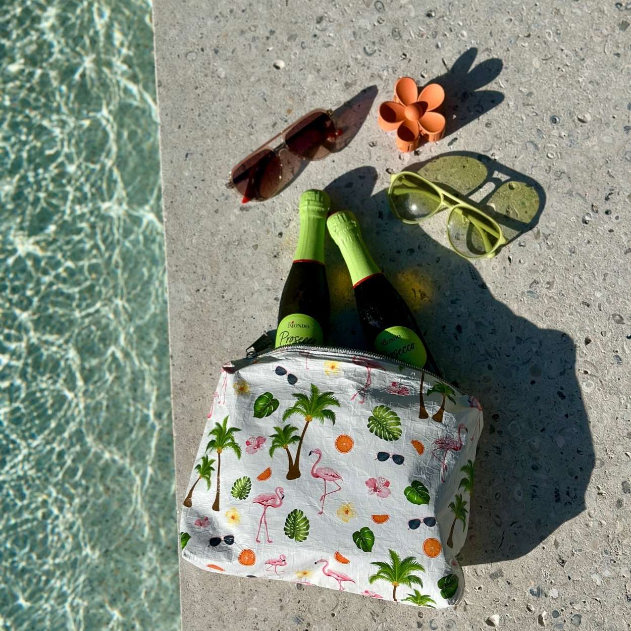 Small bag with flamingo and palm tree pattern, bottles, sunglasses, and a flower on concrete near water.