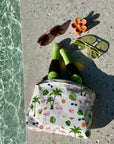 Small bag with flamingo and palm tree pattern, bottles, sunglasses, and a flower on concrete near water.