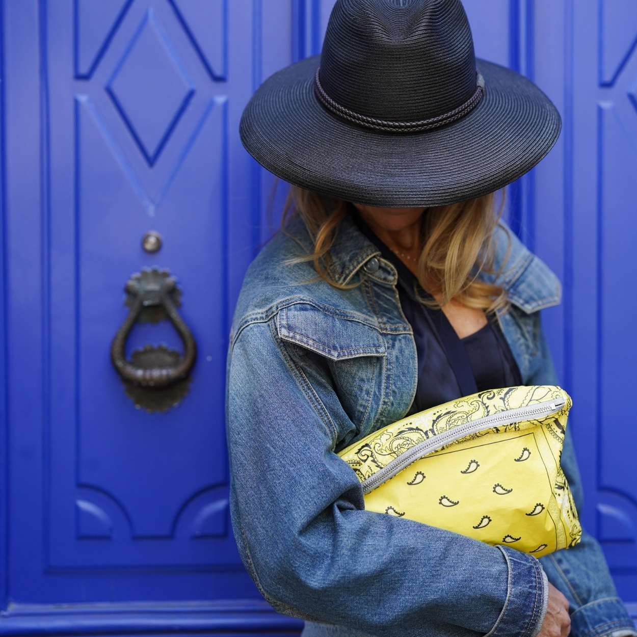 Person wearing a black hat and denim jacket holding a yellow clutch in front of a blue door.