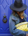 Person wearing a black hat and denim jacket holding a yellow clutch in front of a blue door.